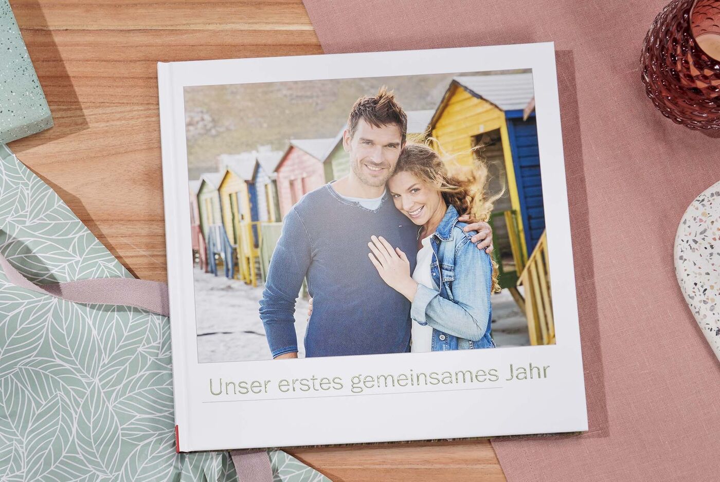Ein Paar steht fröhlich vor bunten Strandhäusern mit einem CEWE FOTOBUCH.
