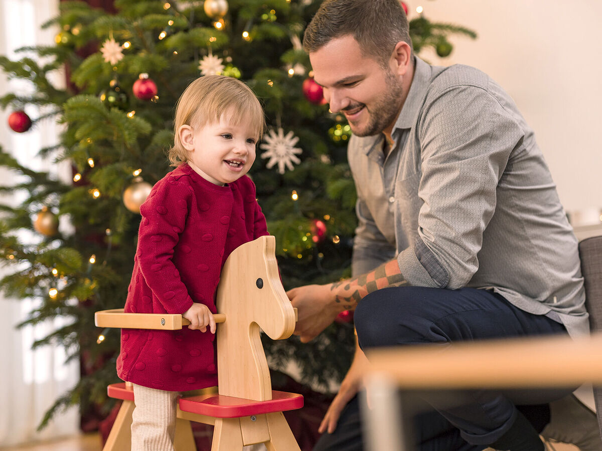 Ein kleines Kind sitzt auf einem Holzspielzeugpferd und schaut den Erwachsenen an, der ein Weihnachtsbaum hinter ihnen dekoriert ist.