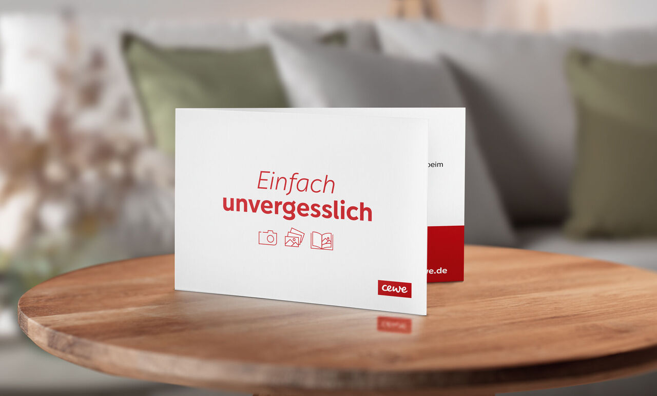 Eine offene Grußkarte mit dem Text "Einfach unvergesslich" von CEWE auf einem Tisch, umgeben von einer gemütlichen Wohnatmosphäre