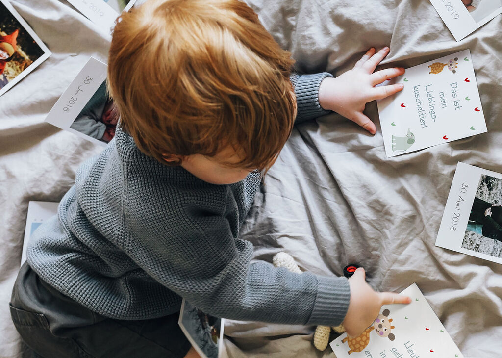 Ein kleiner Junge sitzt auf einem Bett und spielt mit Fotokarten auf einer grauen Decke