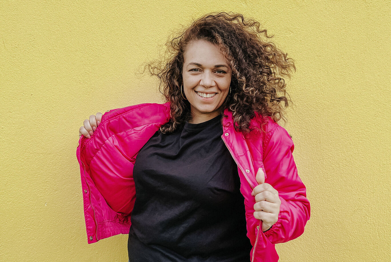 Frau mit lockigem Haar in pinker Jacke vor gelber Wand