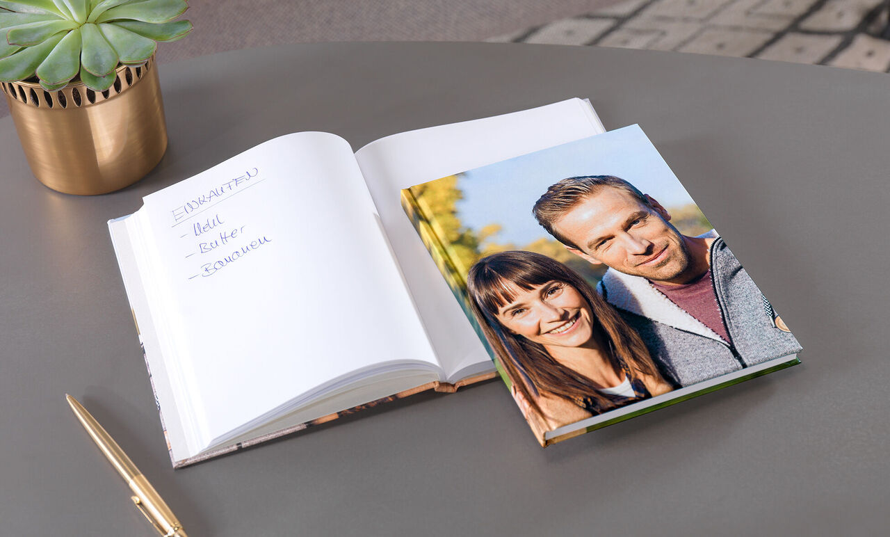 Ein offenes Fotobuch und ein Kalender auf einem Tisch, umgeben von einem grünen Pflanzenarrangement