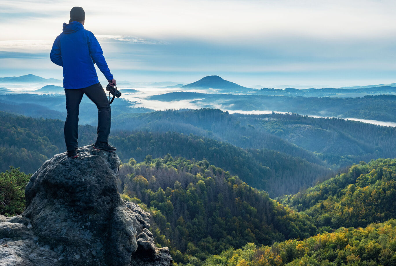 Mann in blauer Jacke steht auf einem Felsen und blickt auf eine malerische Berglandschaft mit Nebel.
