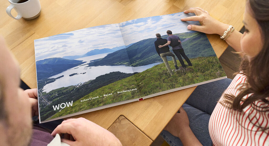 Zwei Personen betrachten ein geöffnetes Fotobuch mit Landschaftsbildern von Schottland und Wales.