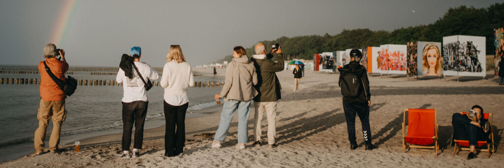 Fotografinnen und Fotografen stehen am Strand und fotografieren einen Regenbogen über der Ostsee. Im Hintergrund ist eine Fotoausstellung.