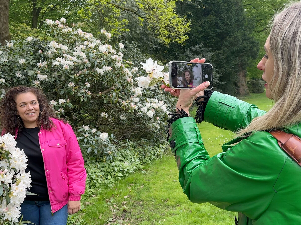 Eine Person in einem pinken Outfit wird von einer anderen Person im grünen Mantel fotografiert, während sie vor blühenden Sträuchern steht.