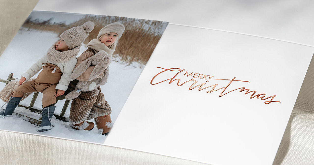 Drei Kinder in winterlicher Kleidung sitzen auf einer Holzbank im Schnee und halten sich um die Schultern. Es ist ein frohes Weihnachtsmotiv.