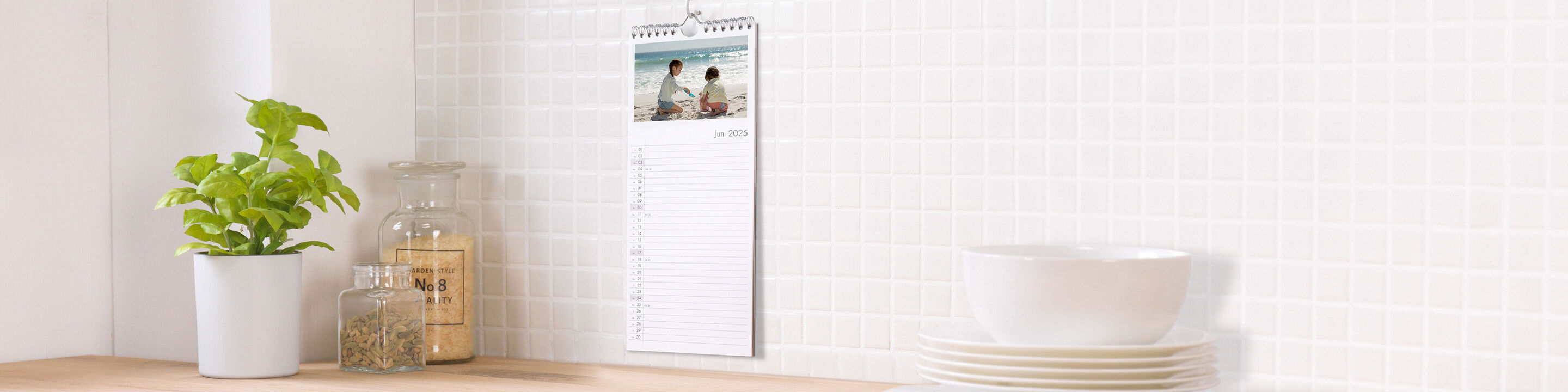 Ein Wandkalender zeigt ein schönes Urlaubsbild mit zwei Personen. Er hängt an einer hellen Wand über einem Holztisch mit einer Pflanze und Geschirr.