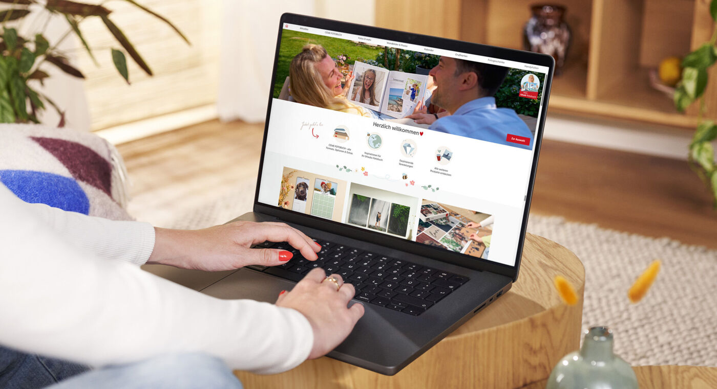 Eine Person sitzt an einem Tisch und betrachtet die CEWE Webseite auf einem Laptop, um kreative Produkte zu gestalten.