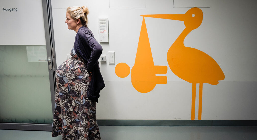 Schwangere Frau geht auf dem Gang einer Klink. An der Wand ein Bild eines Storchs, der Tuch mit Baby im Schnabel trägt.