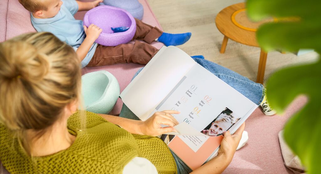 Eine Mutter blättert in einem CEWE Fotobuch auf dem Sofa, während ihr Kind in einem bunten Behälter spielt