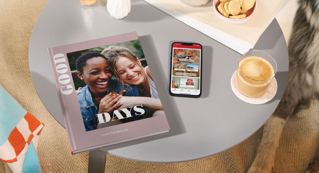 CEWE FOTOBUCH und Smartphone mit CEWE App auf rundem Couchtisch, dazu Kaffee, Kekse und Deko in gemütlicher Atmosphäre.