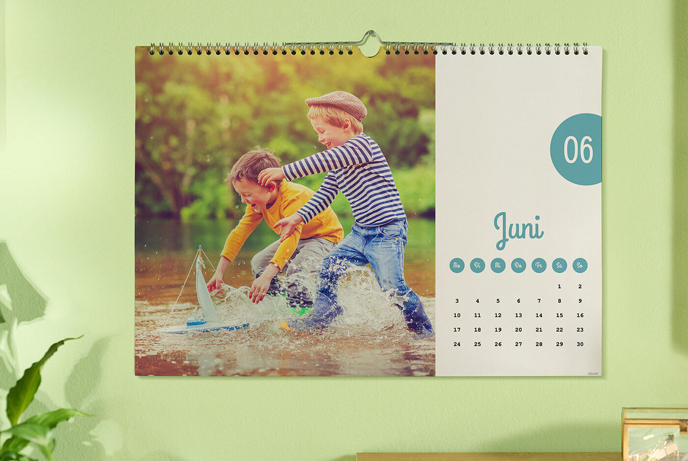 Zwei Kinder spielen im Wasser, halten ein Spielzeugboot. Der Juni-Kalender hängt an der Wand.