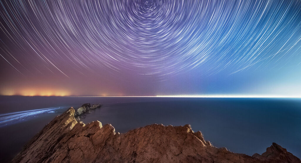 Felsen am Meer bei Nacht, darüber kreisende Sternenspuren am klaren Himmel.