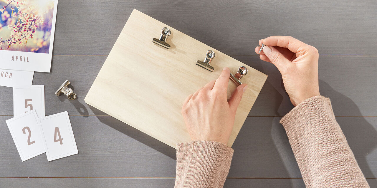 Eine Hand hängt ein Bild mit Clips an eine Holzplatte. Auf dem Tisch liegen weitere Bilder.