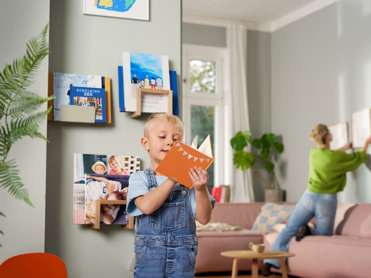 Ein Kind hält eine orange Karte vor dem Gesicht, während im Hintergrund eine Person Bilder an die Wand hängt.