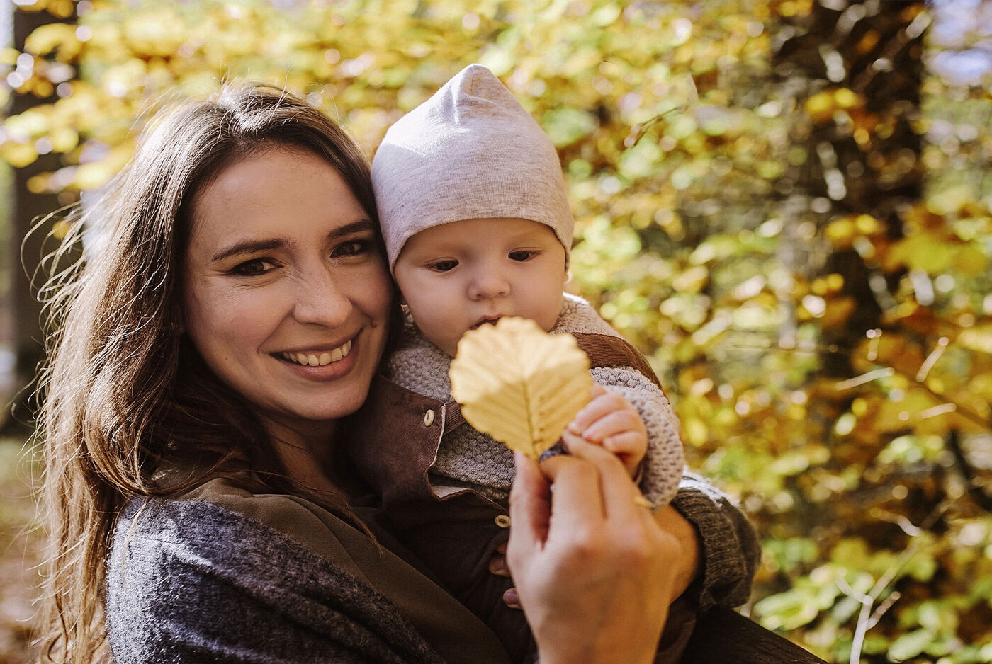 Ein liebevoller Moment im Wald: Erwachsene Person hält ein Kind und ein Blatt in der Hand. Goldene Blätter umgeben sie.