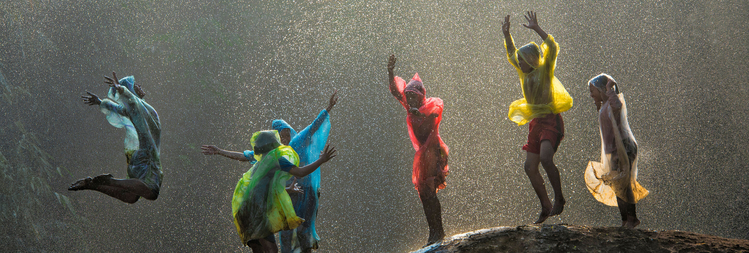Sechs Kinder mit bunten Ponchos hüpfen im Regen auf einem Hügel.