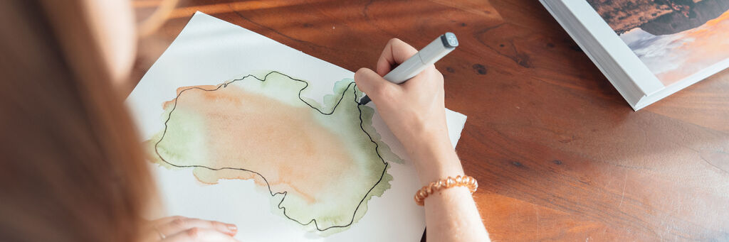Eine Frau zeichnet mit einem blauen Stift eine grüne und orangefarbene Landschaftsskizze auf Papier.
