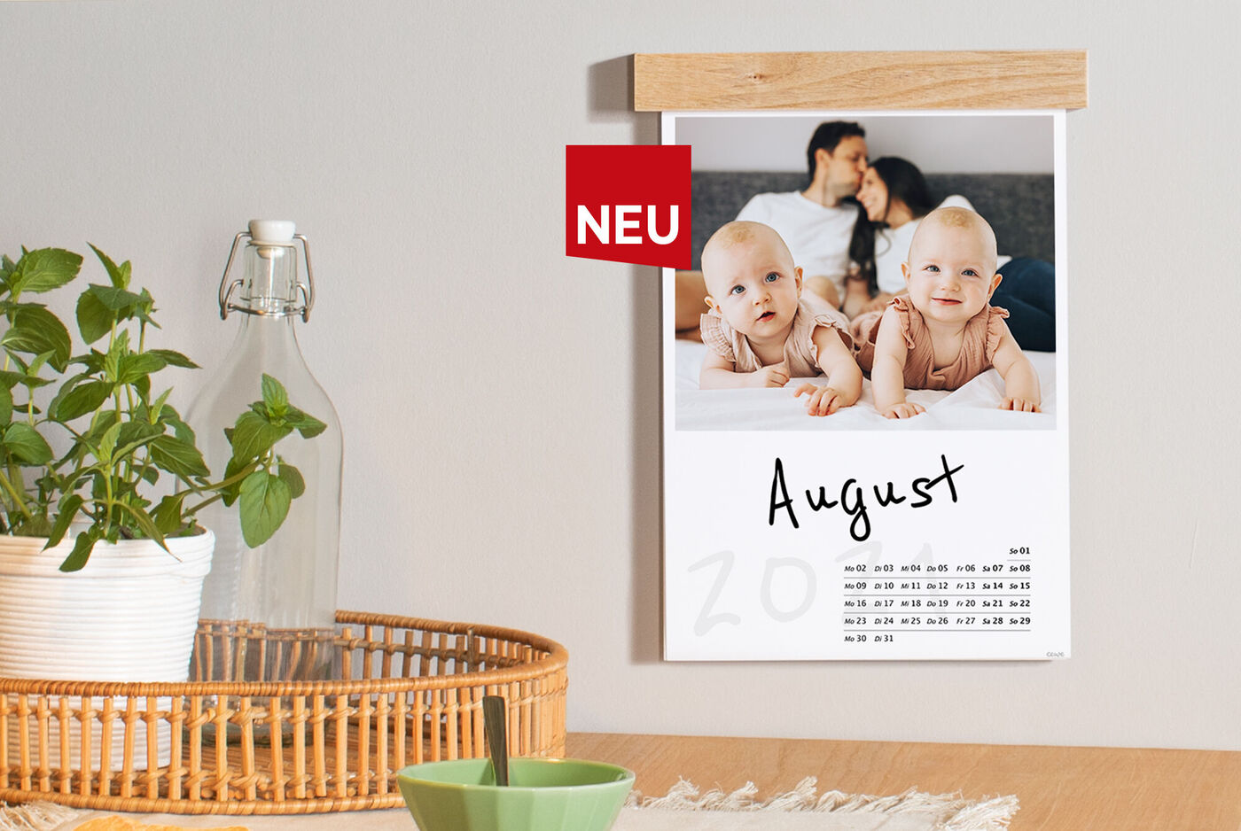 Kalender für August mit zwei Babys, die auf dem Boden spielen. Pflanzen und ein Glas Wasser sind vorhanden.