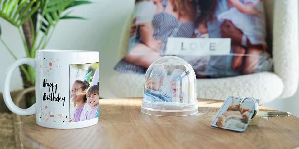 Eine Tasse mit einem Foto und einer Glückwunschbotschaft zum Geburtstag, daneben eine kleine Kuppel mit einem weiteren Bild und ein Schlüsselanhänger mit Fotos, alles auf einem Holztisch.