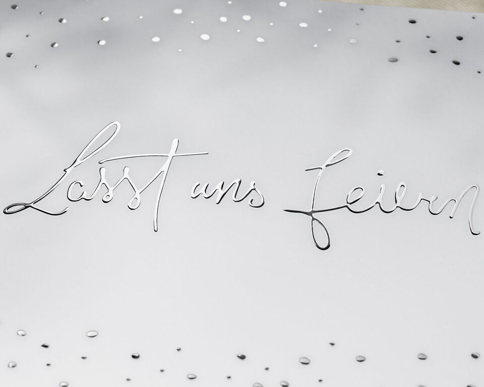 Silberne Schrift mit dem Text 'Lasst uns feiern' auf einem weißen Hintergrund