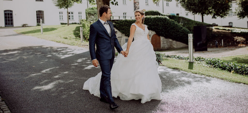 Ein glückliches Paar in Hochzeitskleidung spaziert Hand in Hand auf einem malerischen Weg. Die Atmosphäre ist festlich und voller Freude.
