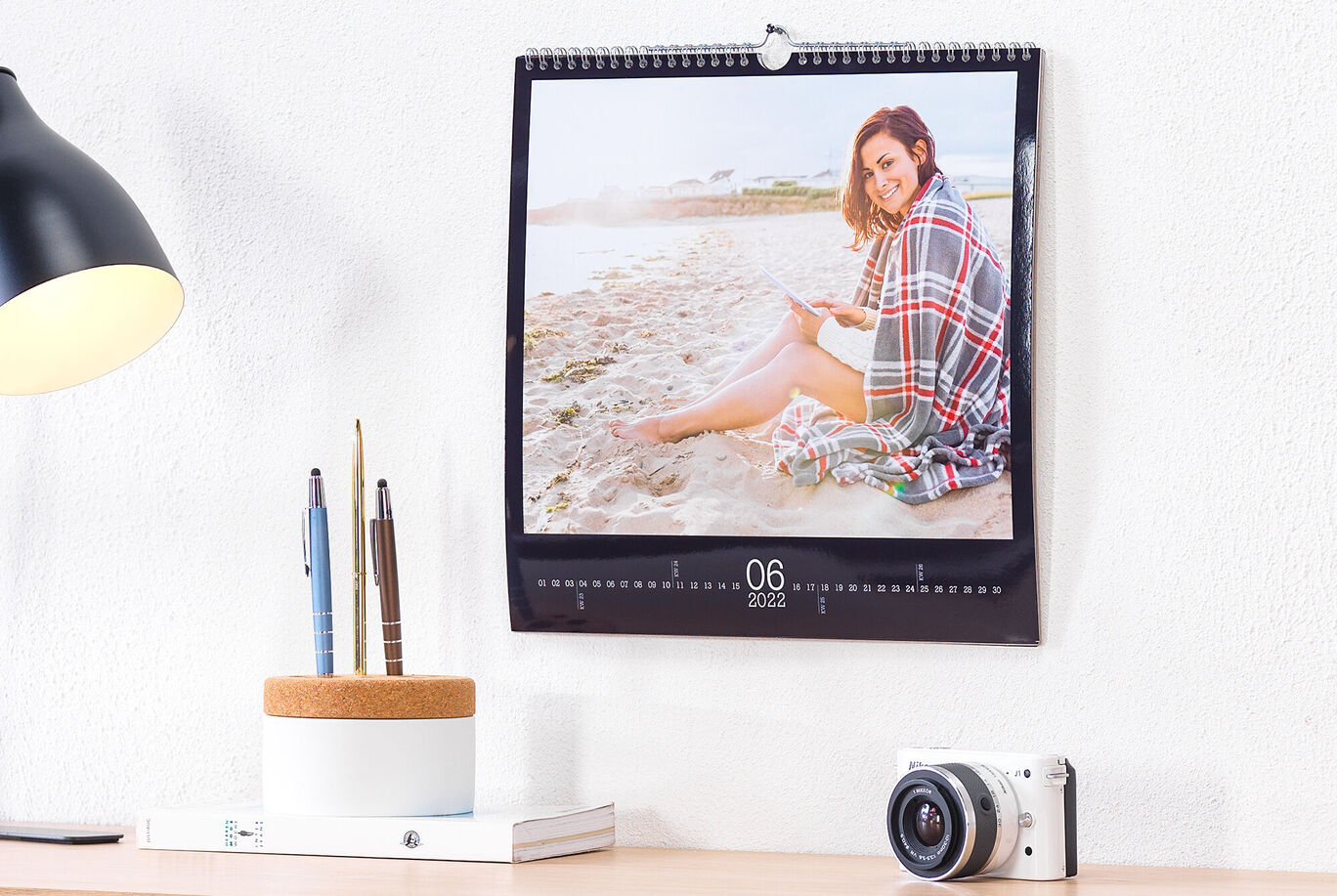 Ein Wandkalender mit einem Bild von einer Person am Strand, die auf einem Handy schaut, umgeben von dekorativen Gegenständen.