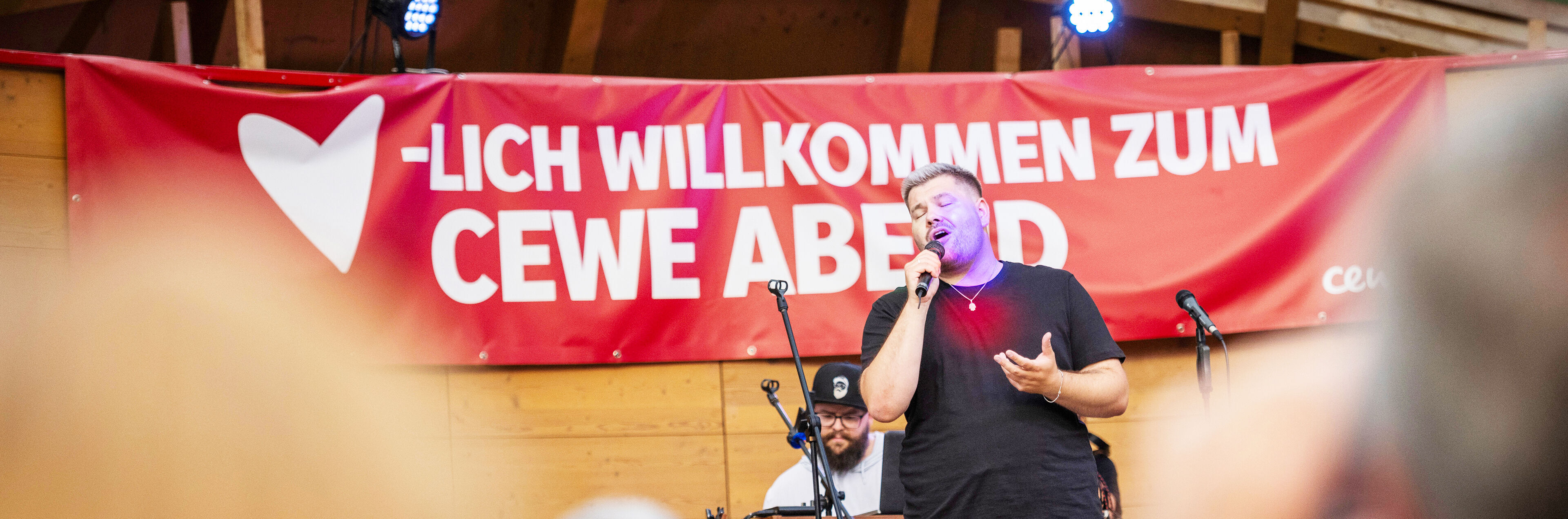 Ein Mann mit einem Mikrofon in der Hand steht vor einem Banner mit der Aufschrift "Herzlich willkommen zum CEWE Abend".