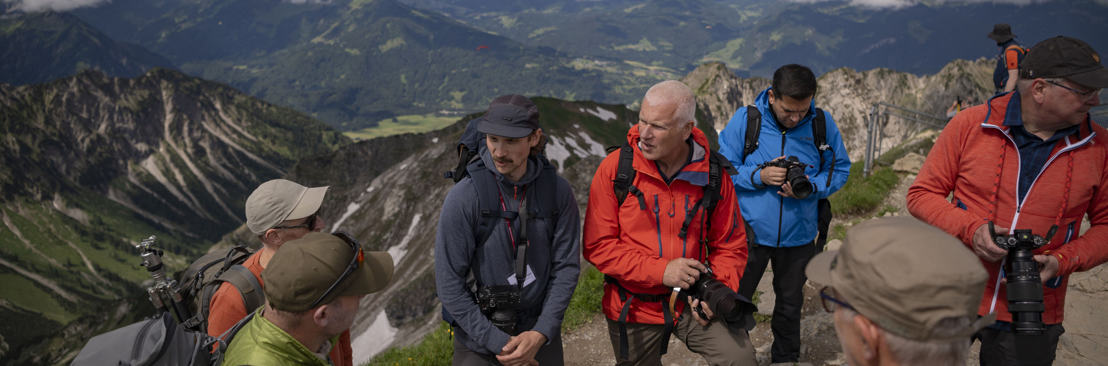 Mehrere Personen in Outdoor-Outfits und mit Kameras in den Händen stehen diskutierend auf einem Berggipfel.