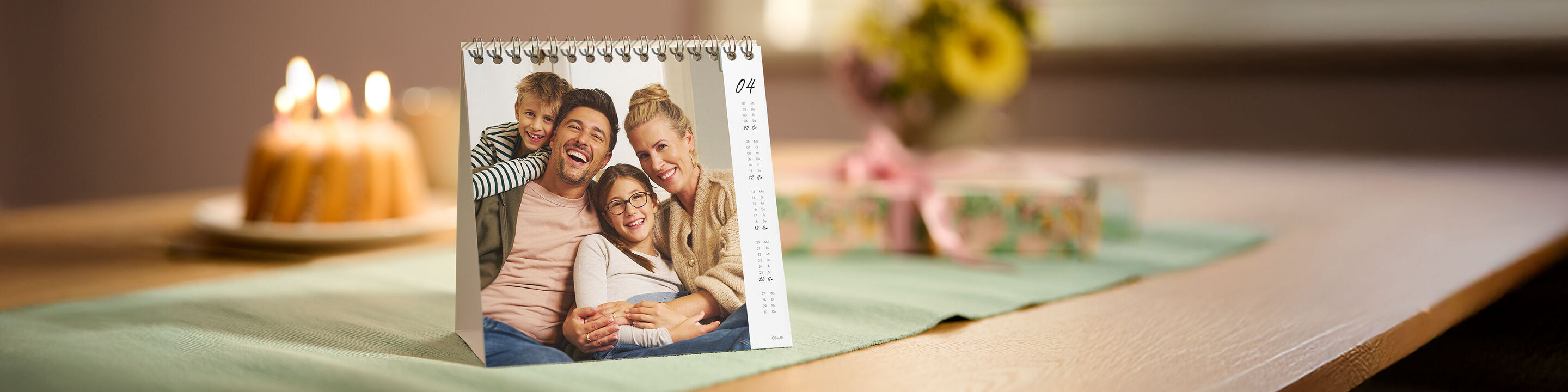 Ein CEWE Fotokalender im Tischformat zeigt im April ein Familien-Porträt und steht dekorativ auf einem festlich gedeckten Tisch.