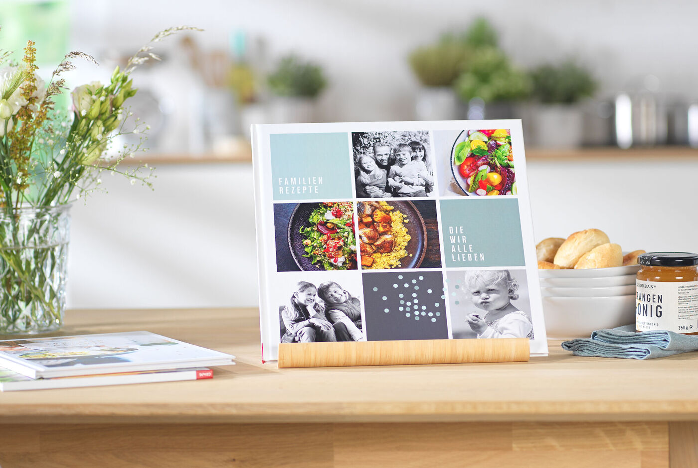 Ein CEWE Fotobuch steht auf einem Holzständer. Es zeigt eine bunte Collage aus Fotos, darunter Bilder von Familie, leckeren Gerichten und schönen Momenten. Im Hintergrund sind frische Blumen und ein Tisch mit Gebäck.