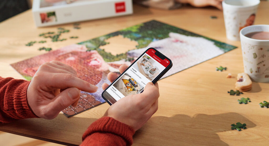 Person hat ein Fotopuzzle vor sich und stöbert auf dem Smartphone in der CEWE App nach Fotogeschenken.