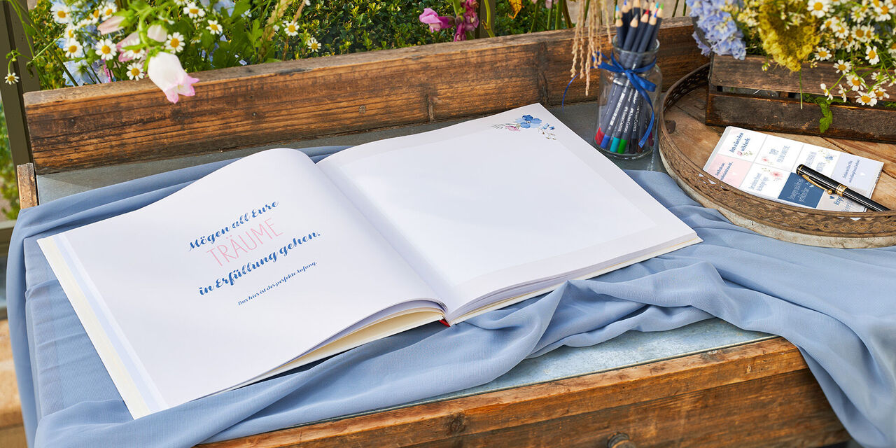 Ein geöffnetes Fotobuch auf einem Holztisch mit bunten Stiften und Blumen im Hintergrund