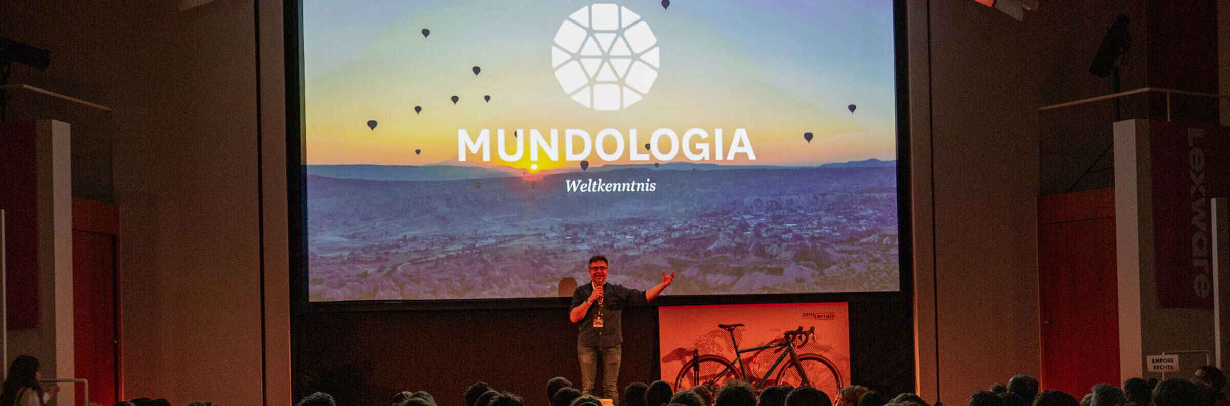 Ein Mann steht auf einer Bühne. Hinter ihm ist eine Leinwand, auf dem das Mundologia-Logo zu sehen ist.