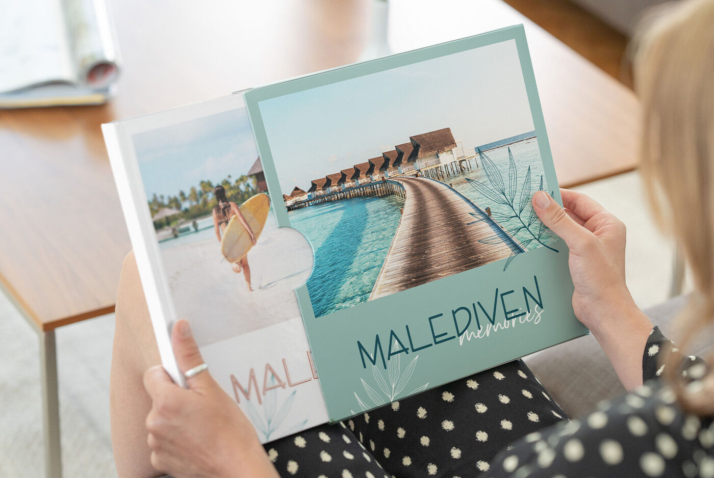 Person betrachtet ein CEWE FOTOBUCH mit Urlaubsbildern von den Malediven, darunter Strandmotive und ein Steg über dem Meer.