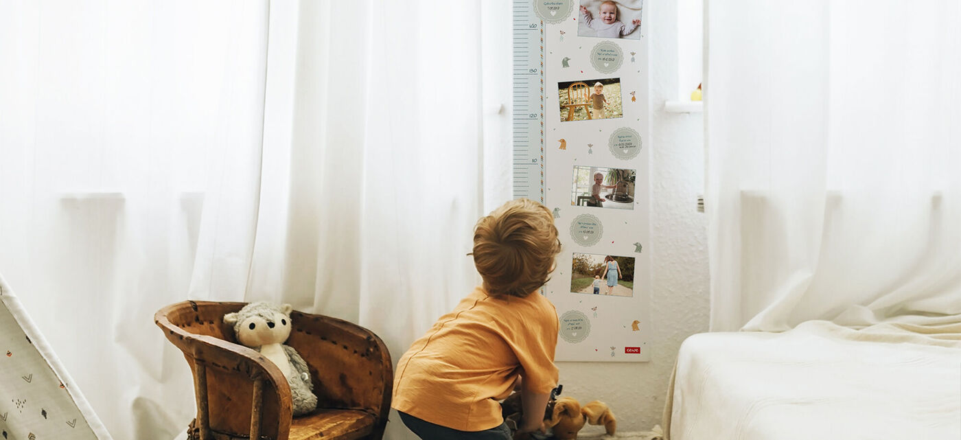 Ein kleiner Junge steht vor einer Wachstumsanzeige und schaut neugierig darauf. Die Wand ist hell und freundlich gestaltet. Neben ihm liegen ein Plüschtier und eine bequeme Couch.
