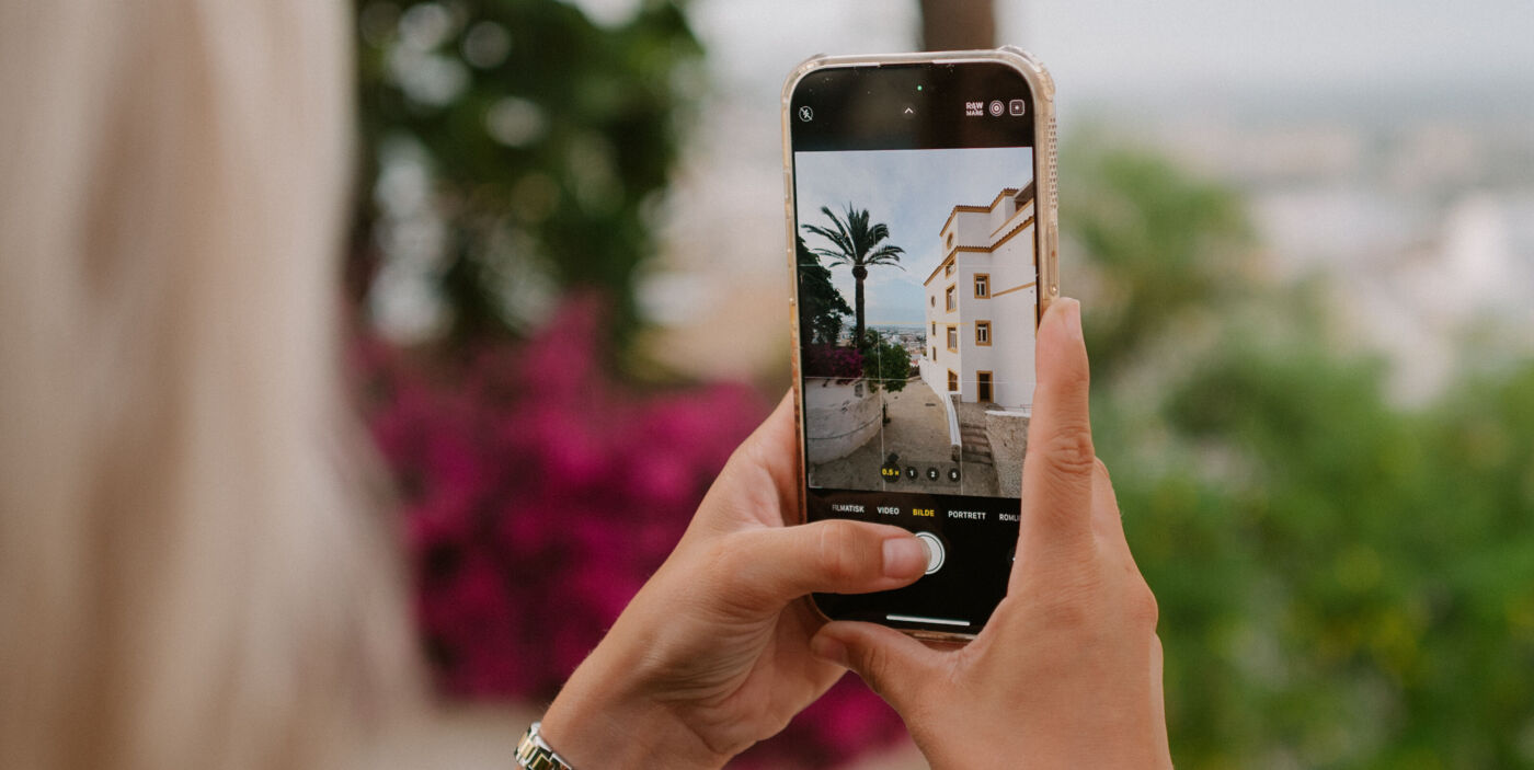 Frau hält Smartphone in Händen, um eine Gasse zu fotografieren. Sie ist gesäumt von einer Mauer, mit Palmen und Gewächsen, und einem Haus.