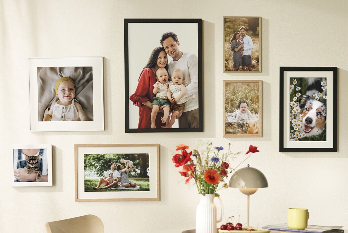 Wand mit CEWE Wandbildern, darunter Familienfotos, Baby, Hund und bunte Blumen in einer Vase auf einem Tisch