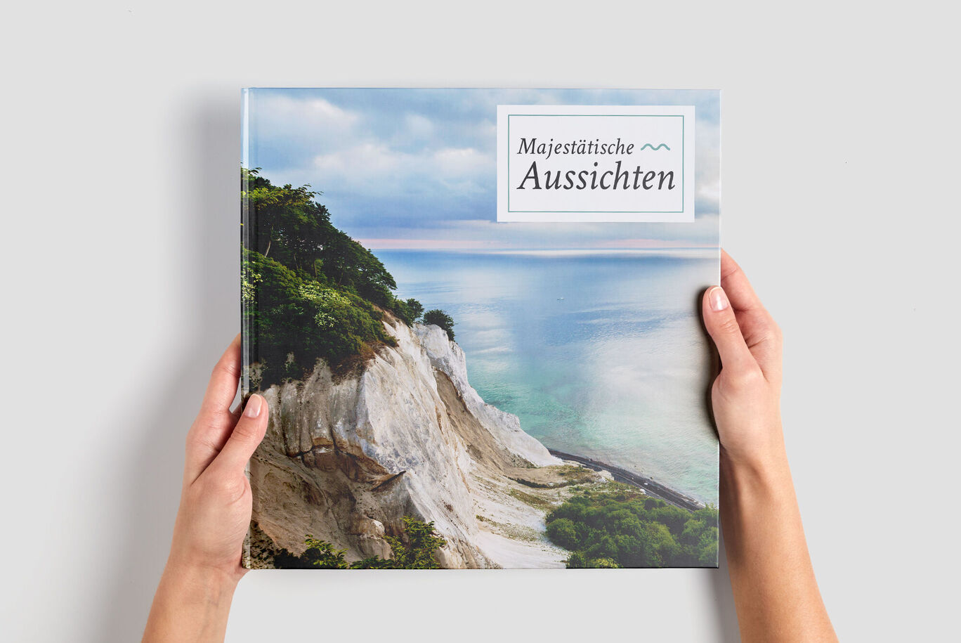Eine Person hält ein CEWE Fotobuch mit dem Titel "Majestatische Aussichten" vor einer schönen Küstenlandschaft.