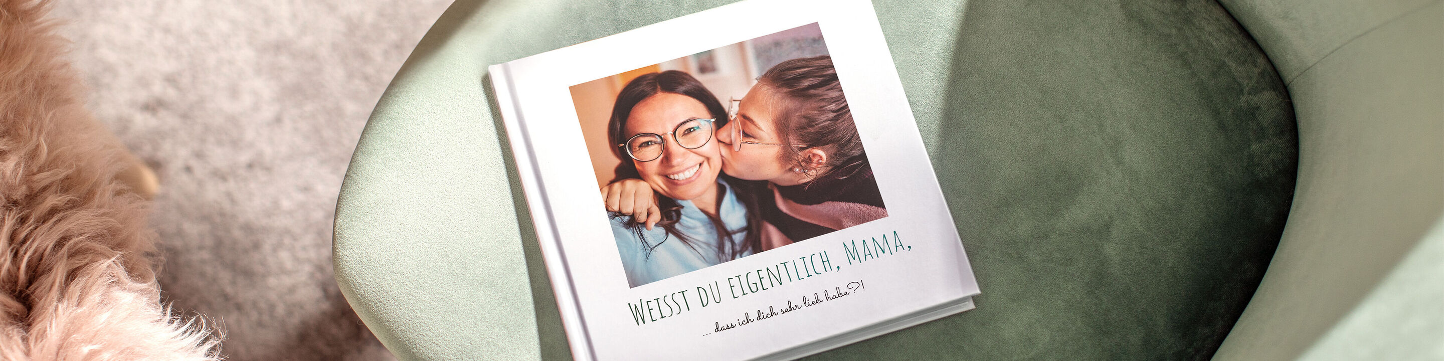 Ein CEWE FOTOBUCH liegt auf einem grünen Sessel. Es zeigt ein liebevolles Bild von zwei Menschen, die sich umarmen