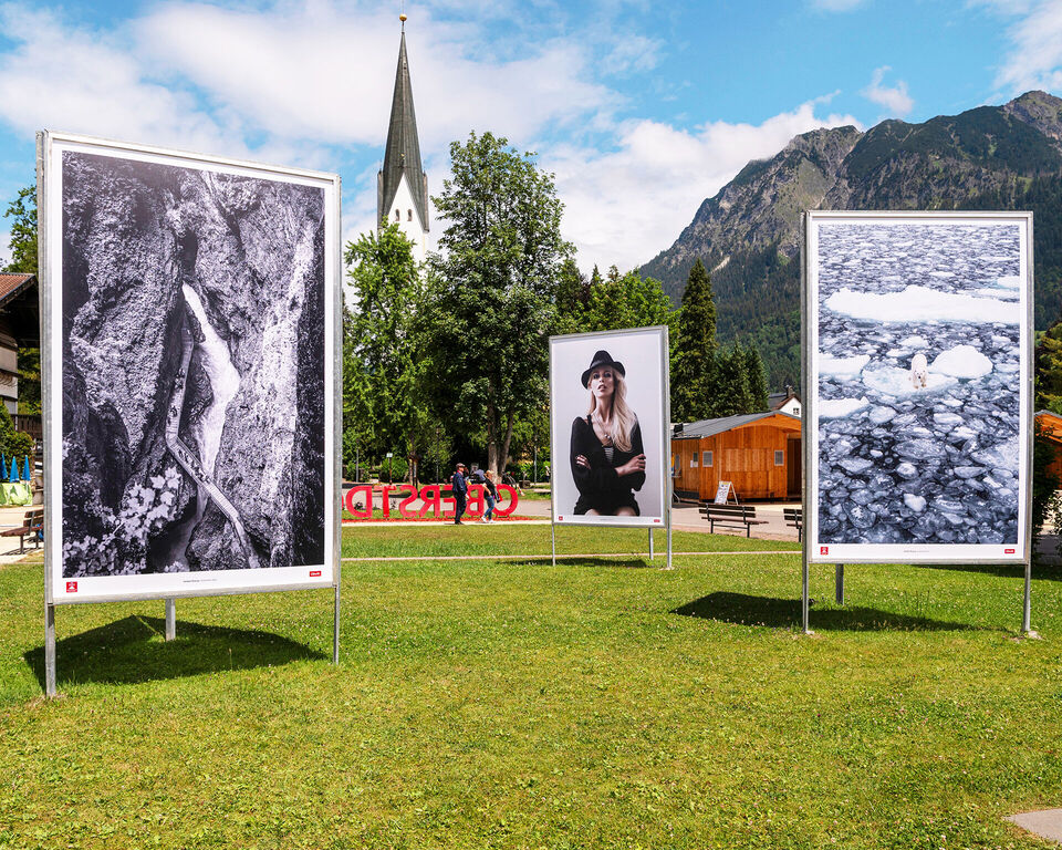 Auf einer Wiese stehen drei große Fotografien. Im Hintergrund ist eine Kirche zu sehen.