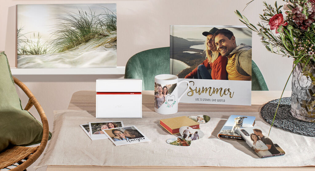 Ein kreatives Arrangement mit Fotoprodukten auf einem Tisch, darunter ein Fotobuch, Tassen und verschiedene Fotos, die Sommererinnerungen zeigen