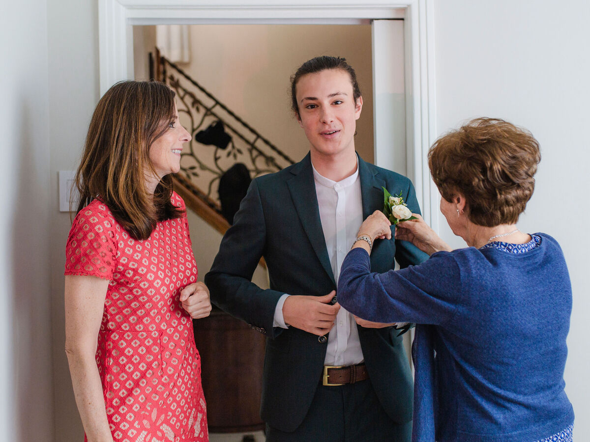 Eine Hochzeitsszene mit einer Frau, die einem Mann eine Blume anheftet, während zwei weitere Personen in feierlicher Kleidung zuschauen.