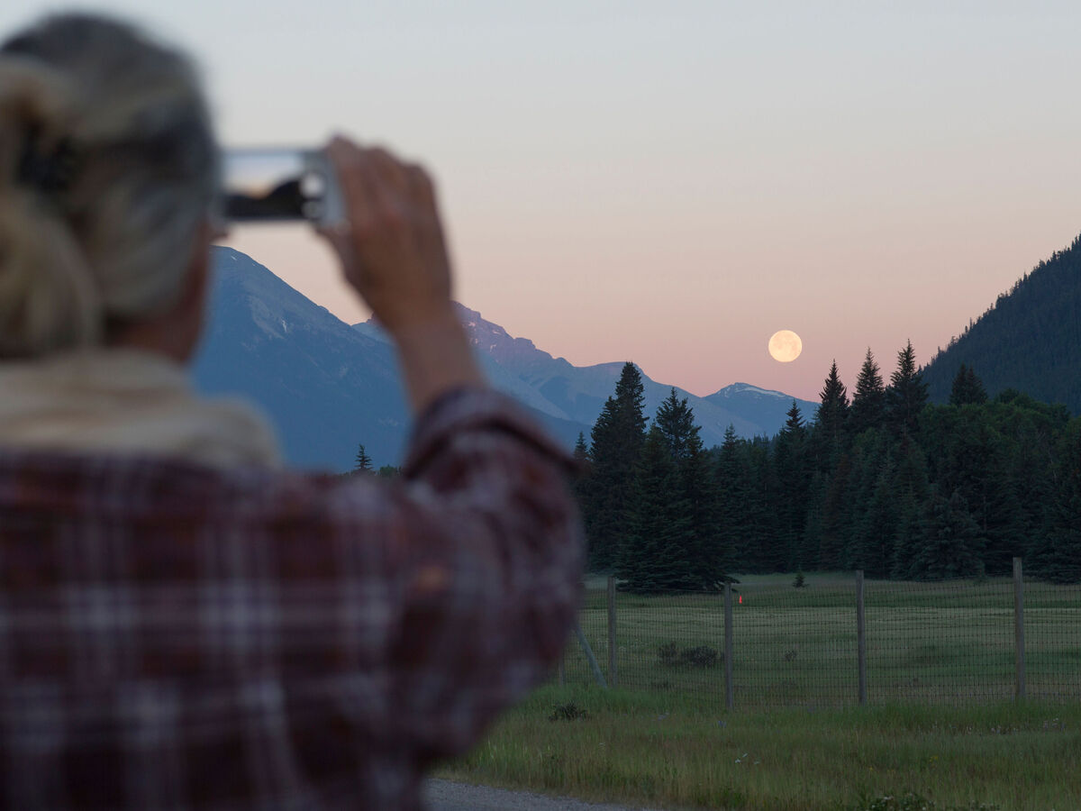 Person fotografiert mit Handy den Vollmond über grüner Wiese und Bergen bei Sonnenaufgang