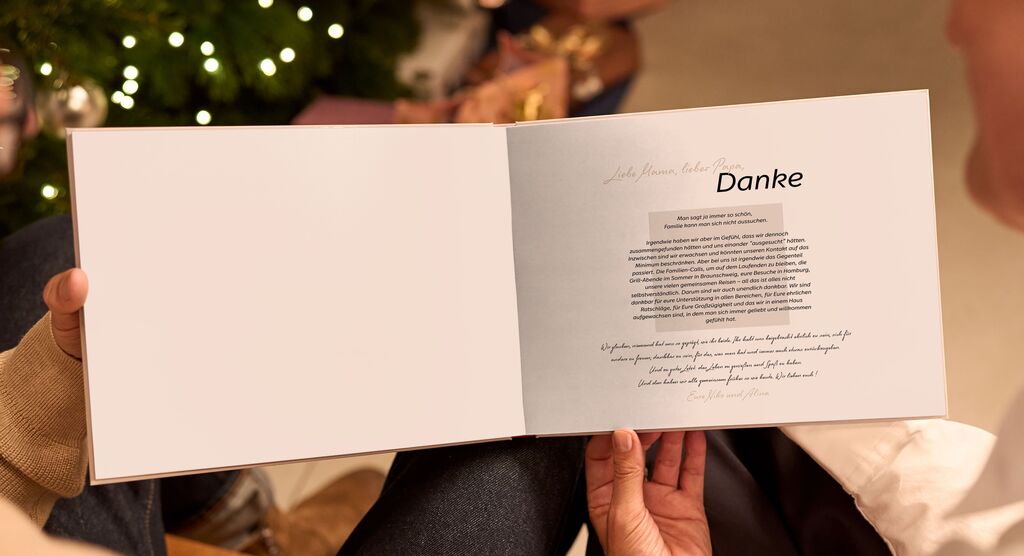 Eine Person hält ein offenes CEWE Fotobuch mit der Aufschrift 'Danke' in Händen. Im Hintergrund sieht man einen geschmückten Weihnachtsbaum.