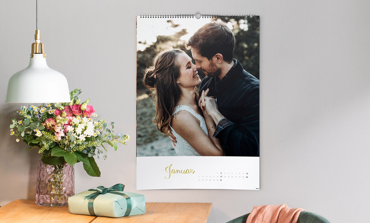 Ein romantischer Wandkalender für den Monat Januar mit einem liebevollen Paar, das sich umarmt. Der Blumenstrauß und das Geschenk auf dem Tisch schaffen eine angenehme Atmosphäre.