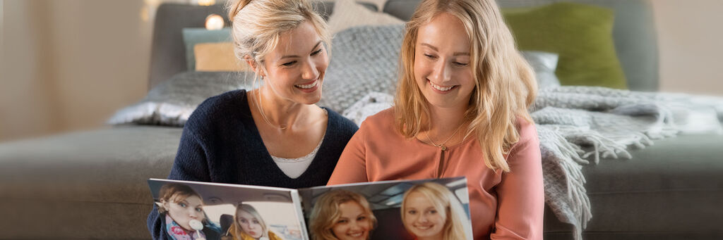 Zwei Frauen betrachten gemeinsam ein CEWE Fotobuch auf dem Sofa und genießen die schönen Erinnerungen.