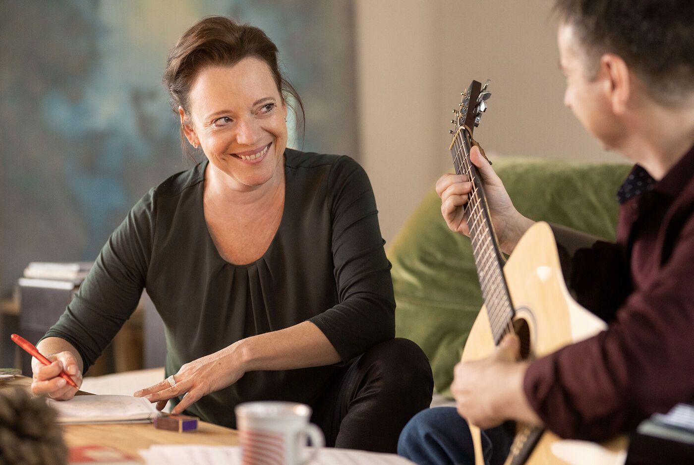 Zwei Personen beim gemeinsamen Musizieren in einem gemütlichen Raum. Eine Person hält eine Gitarre, die andere schreibt Notizen.