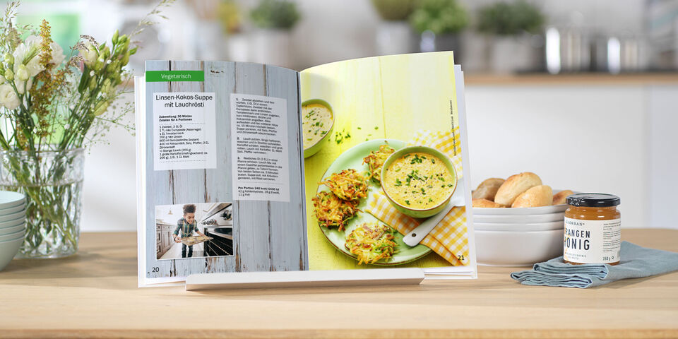 Offenes Kochbuch mit Rezepten für köstliche Gerichte auf einem Tisch, umgeben von Zutaten und frischen Blumen
